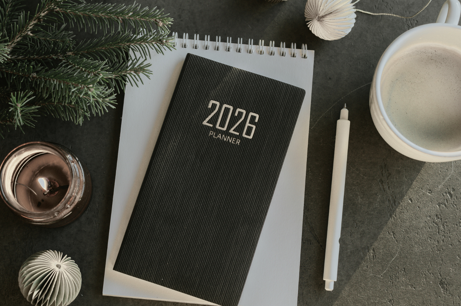 Musta 2026 Planner muistikirja, joulukuusen oksia, kynttilä ja kahvikuppi harmaalla kivipöydällä.